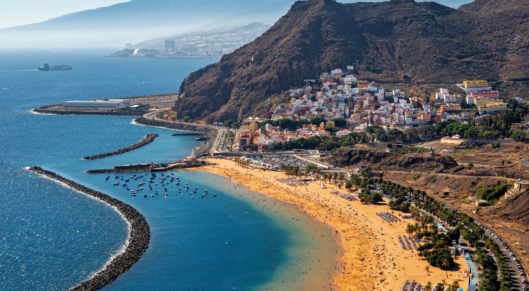 Playa de las Teresitas seen from the hillside above San Andres, its breakwater and palms curving round the bay, with Teide in the distance